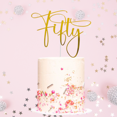 White cake with a gold "Fifty" topper and colourful sprinkles against a pink background.