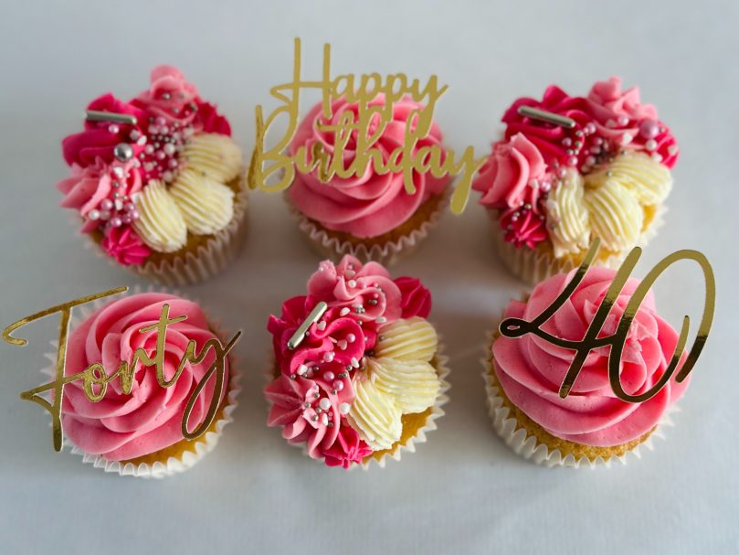 Pink frosted cupcakes decorated with gold birthday toppers and white fruit accents.