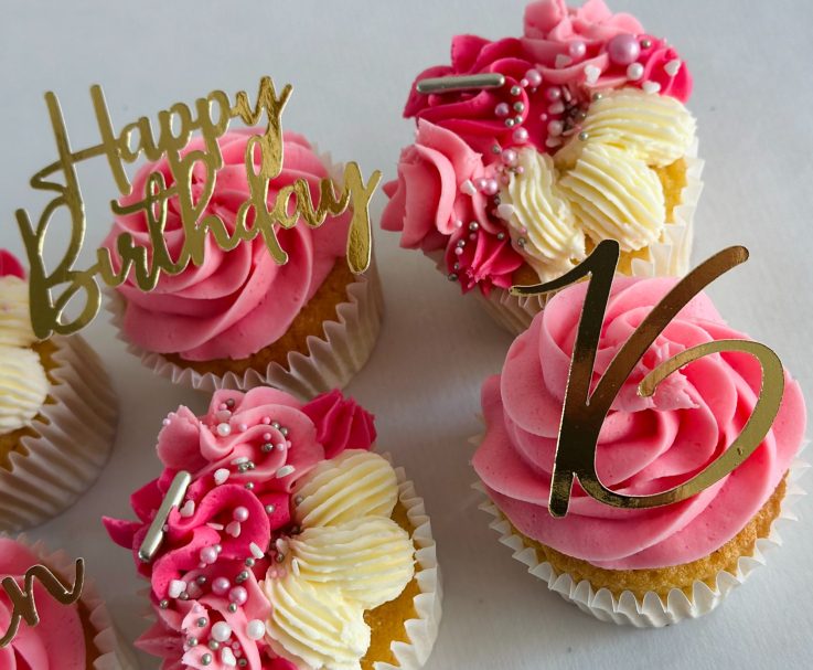 Six celebratory cupcakes with pink frosting and golden "Happy Birthday" toppers.